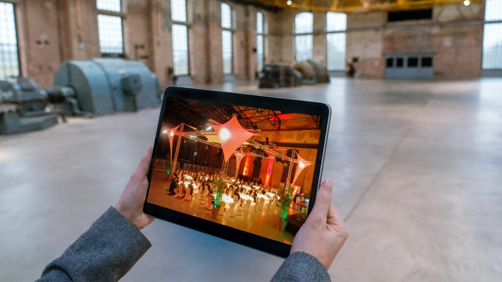 Ein Foto von einem Tablet das von einer Person gehalten wird. Auf dem Tablet ist eine belebte Abendveranstaltung in der Halle zu sehen.